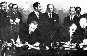 Two men signing an agreement, with other men standing behind them